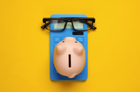 blue calculator with pink piggy bank on top of it and some glasses on a yellow background.