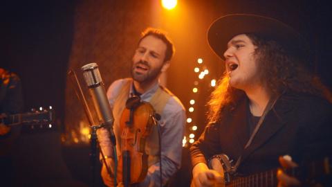 Photo of two musicians performing a song with string instruments and singing under stage lights.