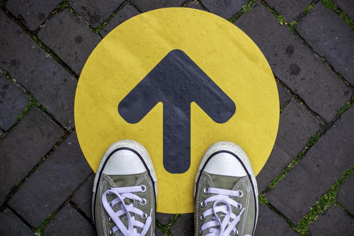 two feet in shoes facing forward standing on top of an arrow indicating forward movement.