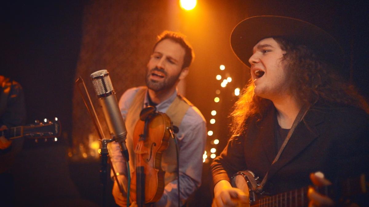 Photo of two musicians performing a song with string instruments and singing under stage lights.